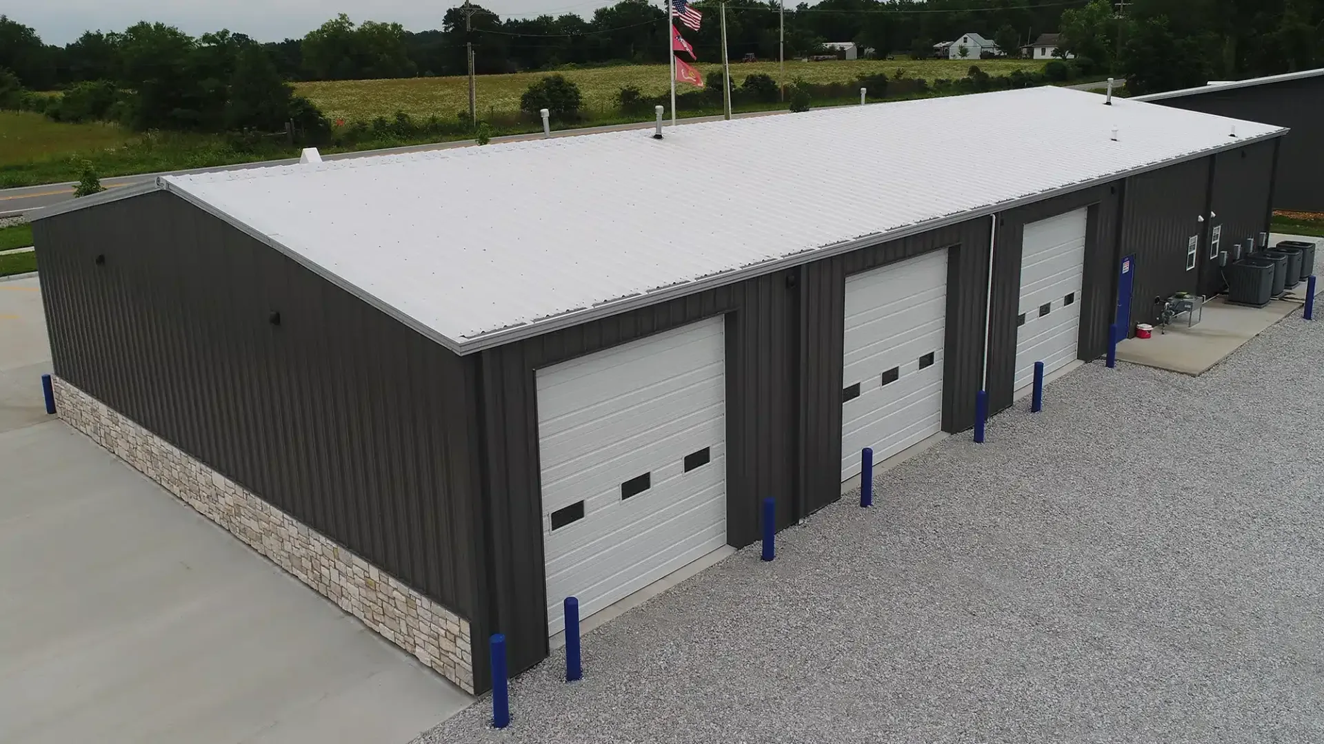 Metal building with three garage doors, gray exterior, white roof, and gravel parking area.