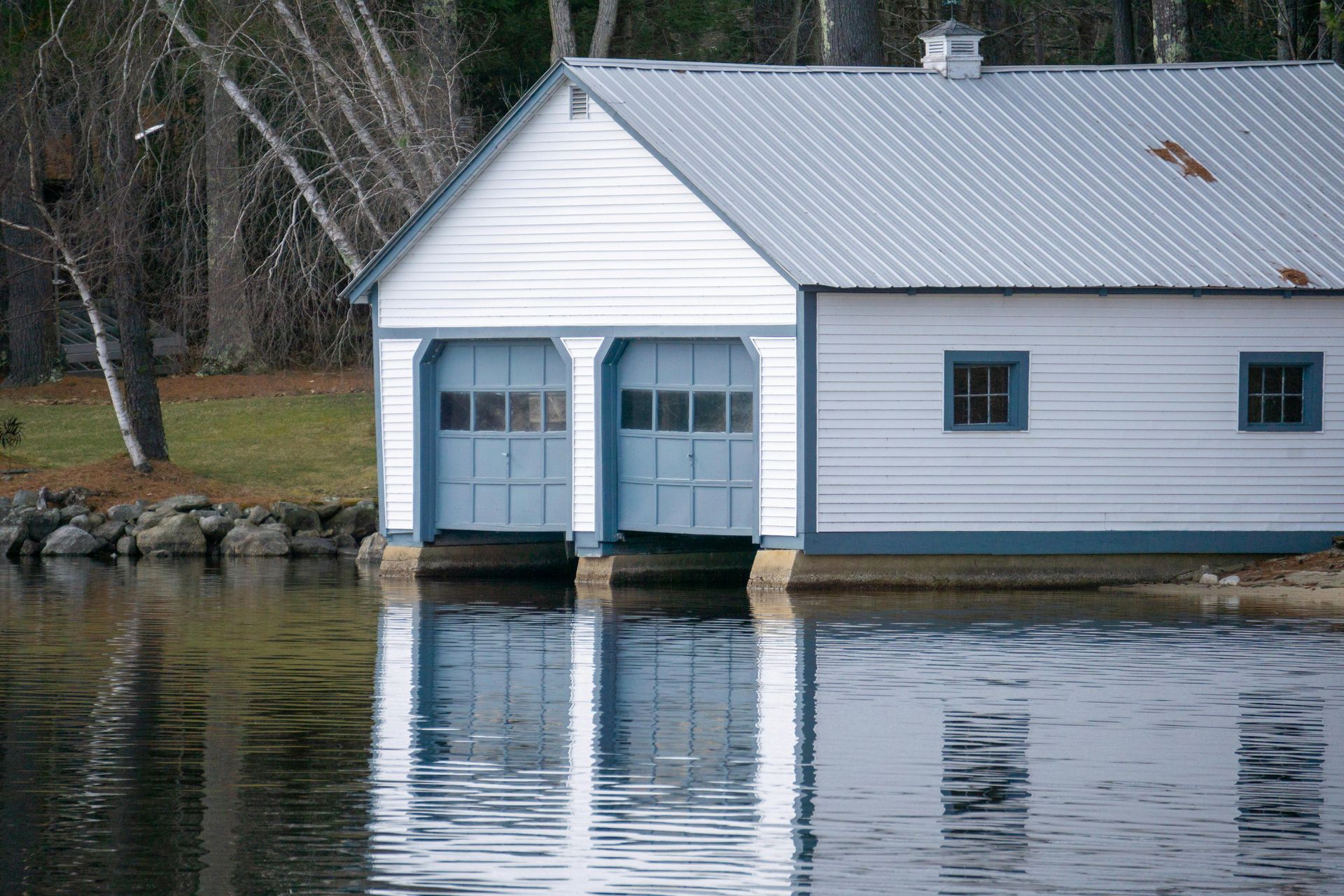 White boathouse on a lake with blue doors and a corrugated metal roof. New construction.