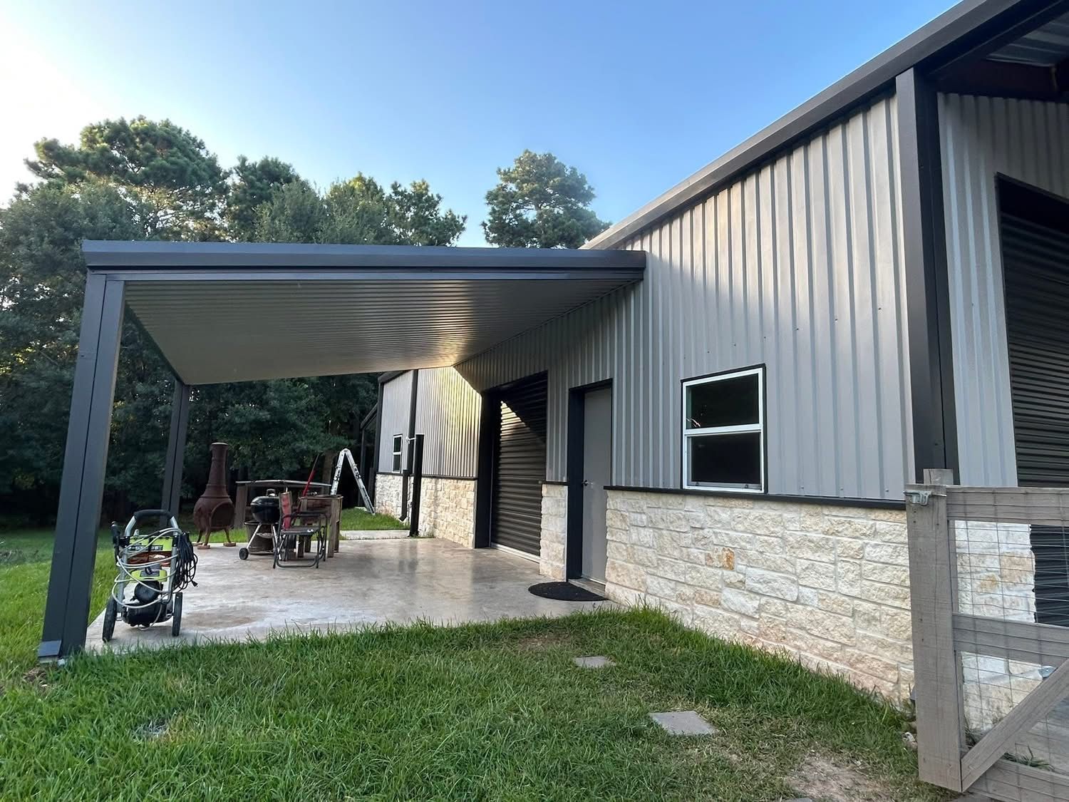 A modern metal building, covered patio, gray siding, stone veneer, and a dark patio cover. New construction,