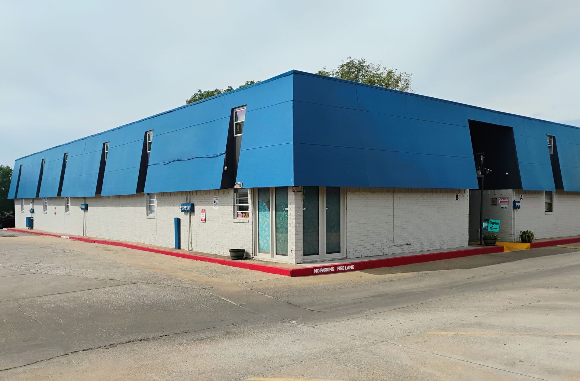 Blue-roofed, two-story commercial building with light gray walls and a red curb, on a paved lot.