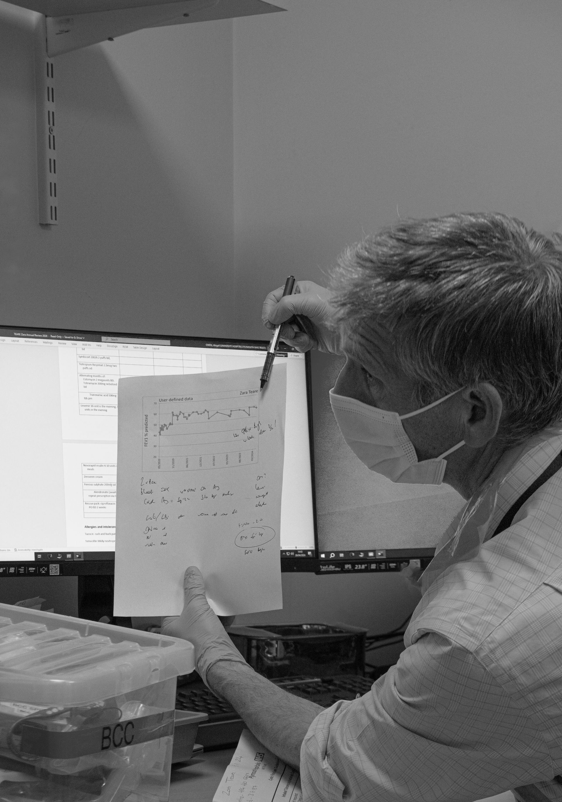 A man wearing a mask is holding a piece of paper in front of a computer monitor.