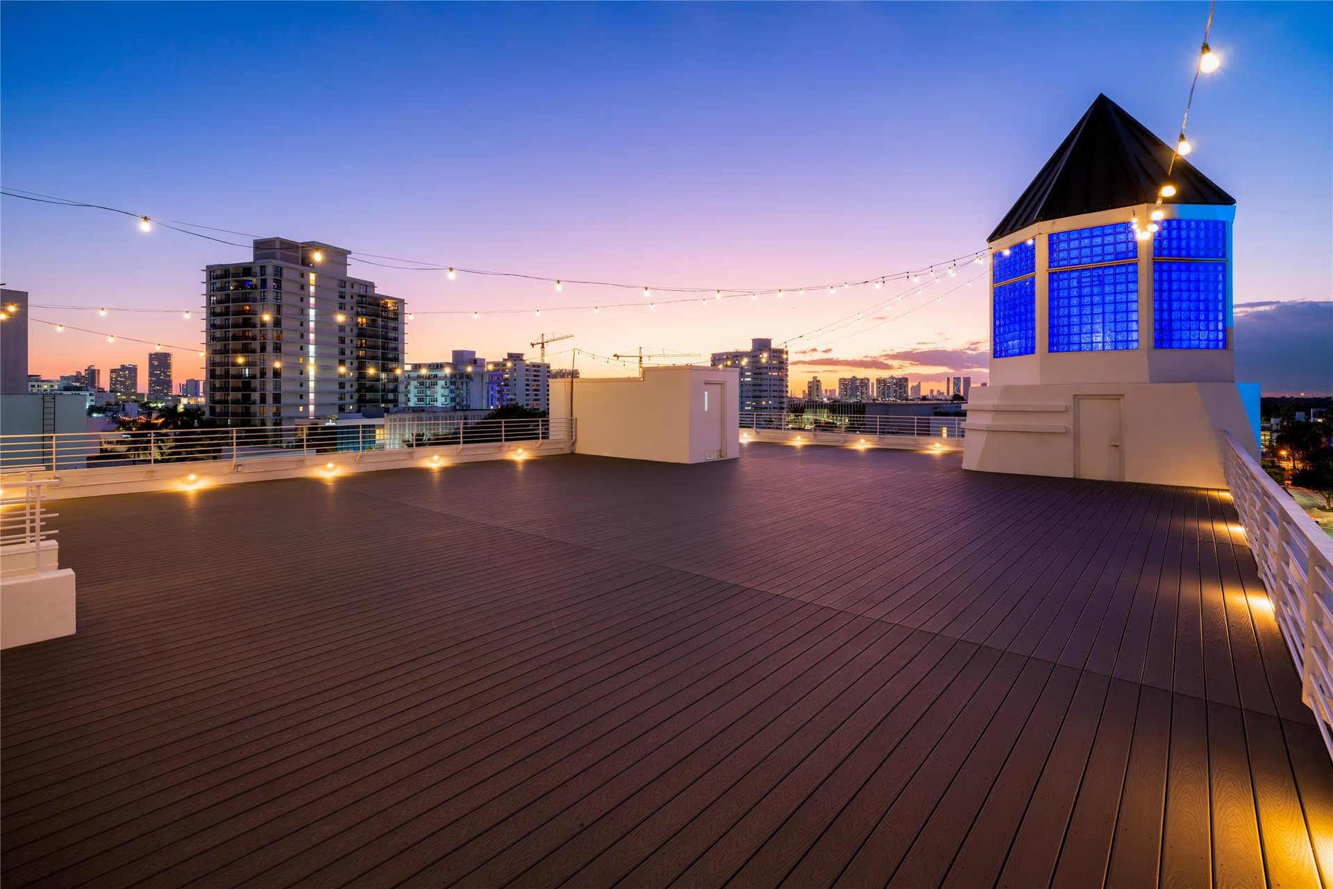 Rooftop deck at sunset with string lights, city buildings, and a blue-lit tower