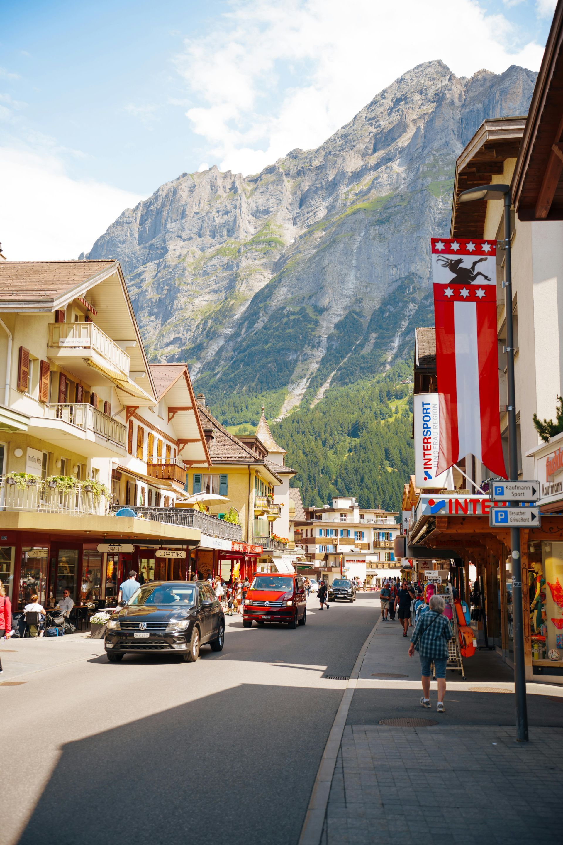 A busy mountain town street with shops, cars, pedestrians, and steep rocky peaks in the background