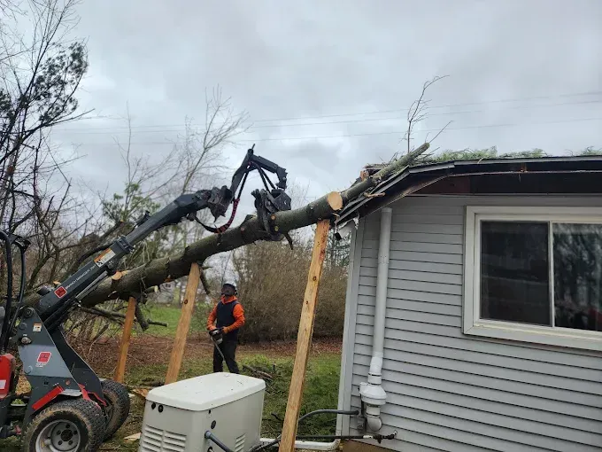 A worker uses heavy machinery to lift a fallen tree away from the damaged roof of a house.