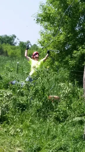 A person in a red helmet and bright yellow shirt holds a cable suspended in a lush, green, overgrown outdoor area.
