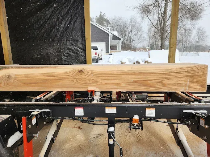A large, freshly milled wooden beam sits on a portable sawmill outdoors in a snowy landscape.