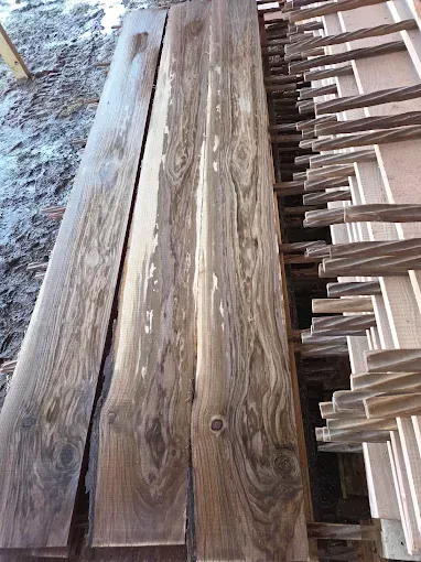 Rough-sawn walnut lumber boards stacked outdoors next to a pile of wooden fence stakes.