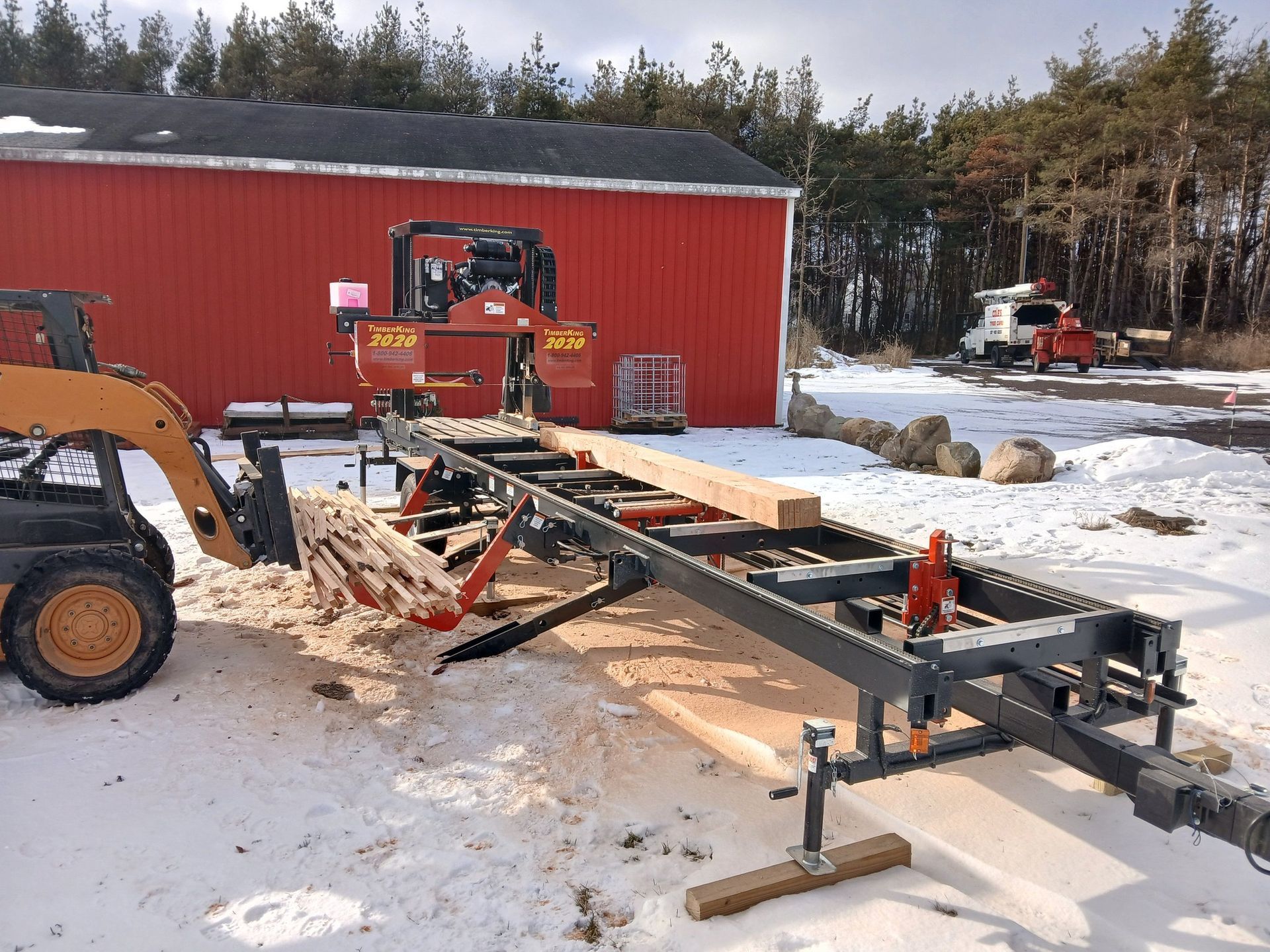 A portable sawmill cutting a large wooden beam, with a skid steer nearby, set against a red barn in a snowy landscape.