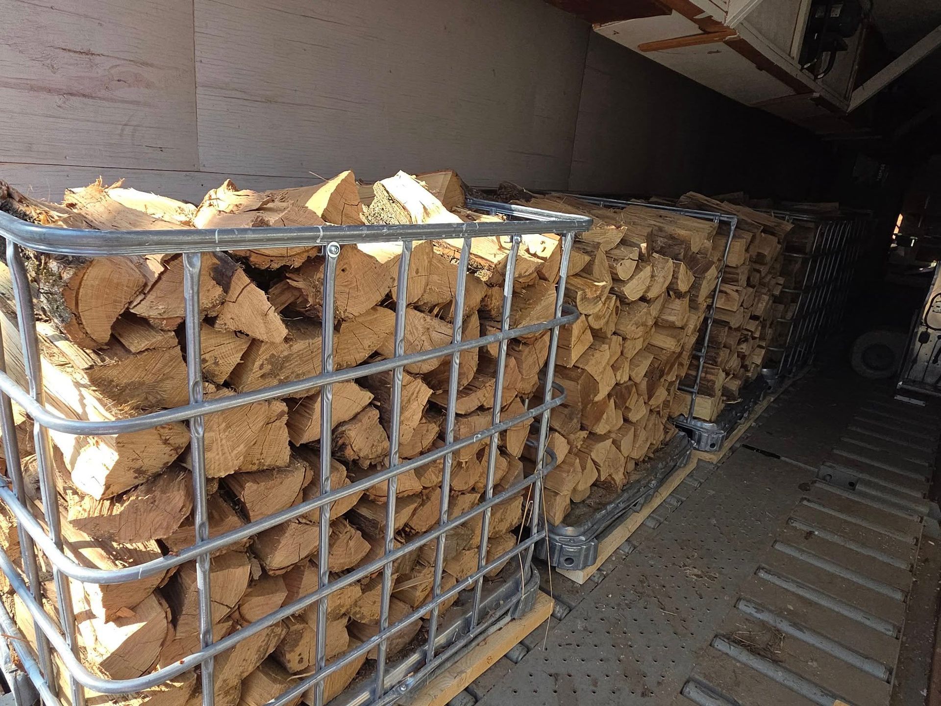 Stacked firewood in metal wire cages resting on pallets inside a transport trailer.