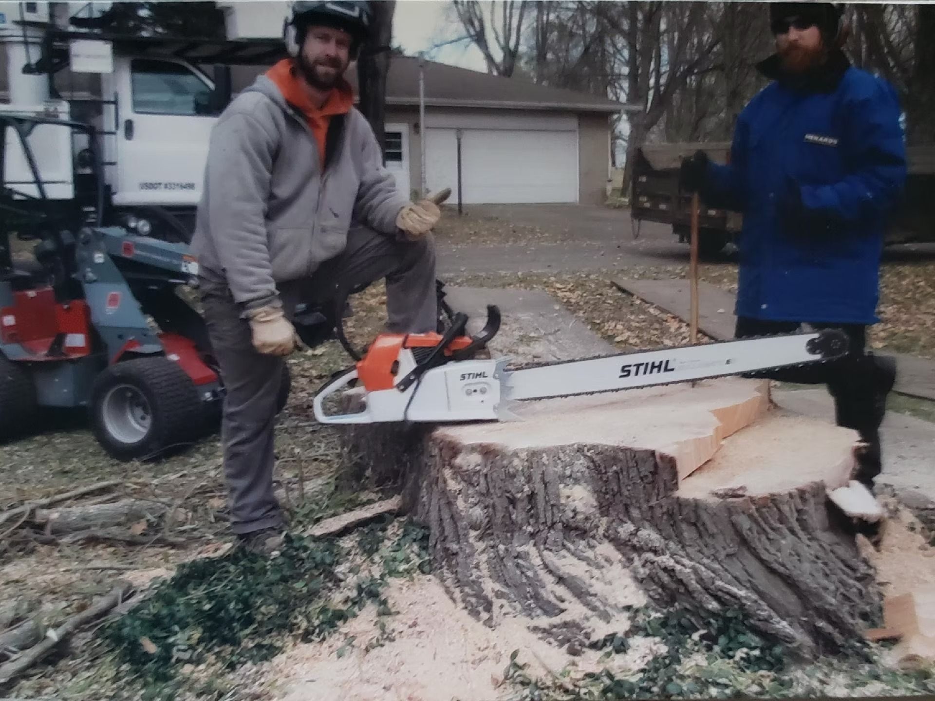 Two workers stand by a large tree stump with a Stihl chainsaw, outdoors near a parked utility truck.
