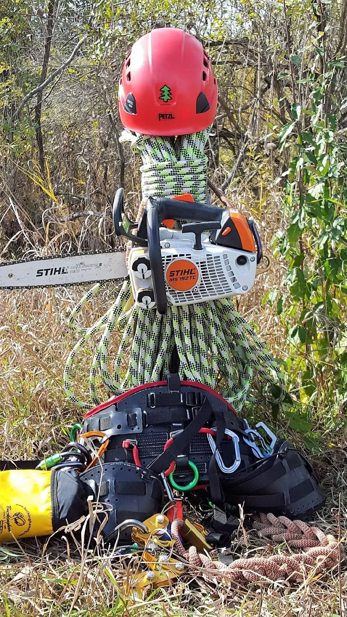 A red climbing helmet rests on a coiled climbing rope and chainsaw, set against a blurred background of brush.