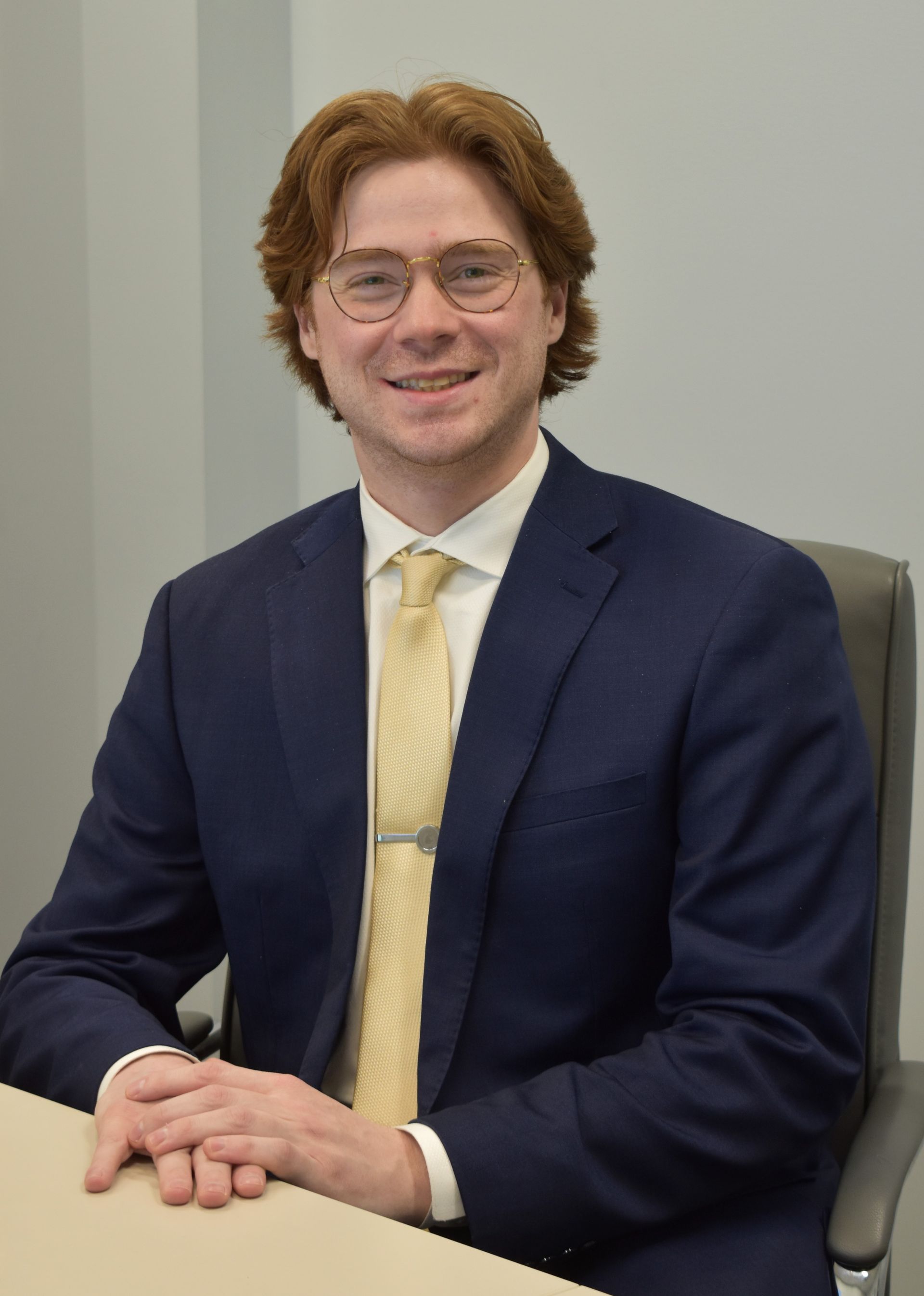 Tyler Brown. A man sits at a desk, smiling, wearing glasses, and wearing a dark colored suit with a cream tie.