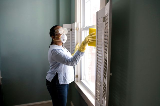 Person wearing safety glasses, mask, and gloves cleans window shutters with a sponge.