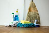 Cleaning supplies on wooden floor: mop, broom, spray bottle, sponge, brush, and bottles.