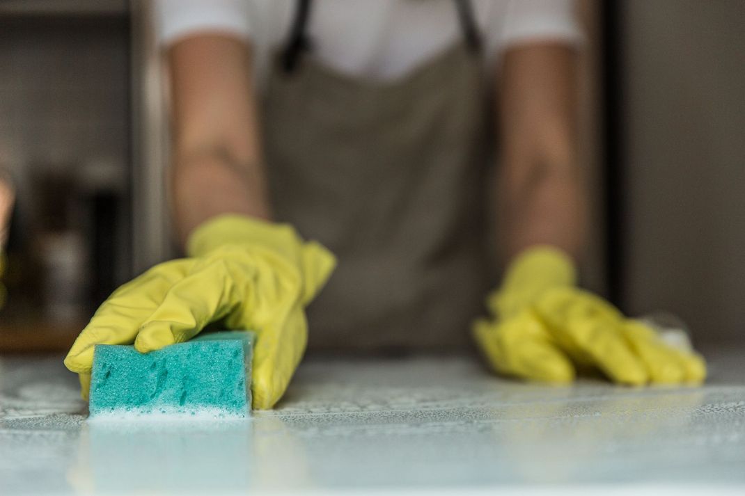 Person wearing yellow gloves wiping a counter with a blue sponge.