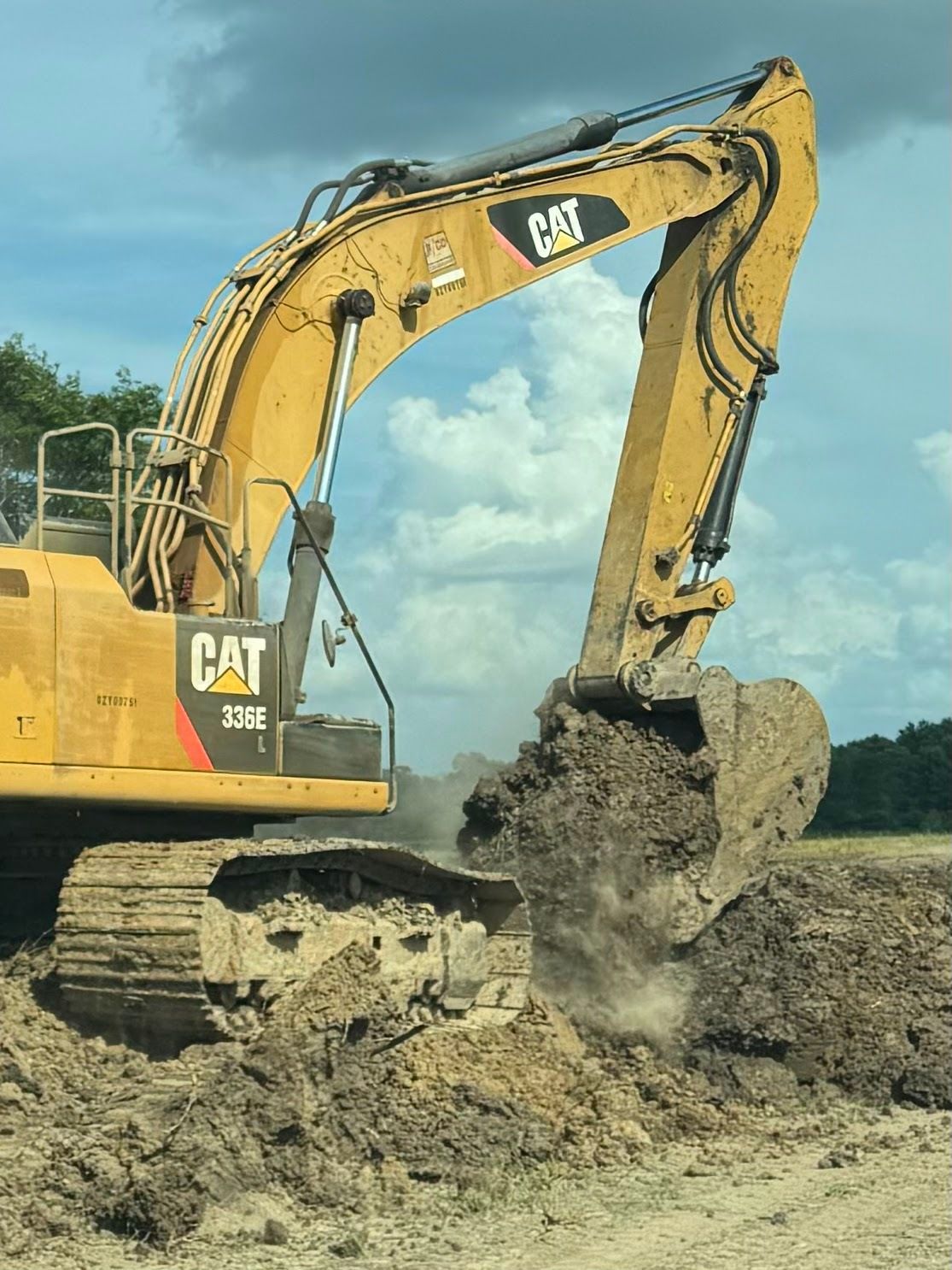 Yellow excavator bucket next to a field with green crops and a cloudy sky.