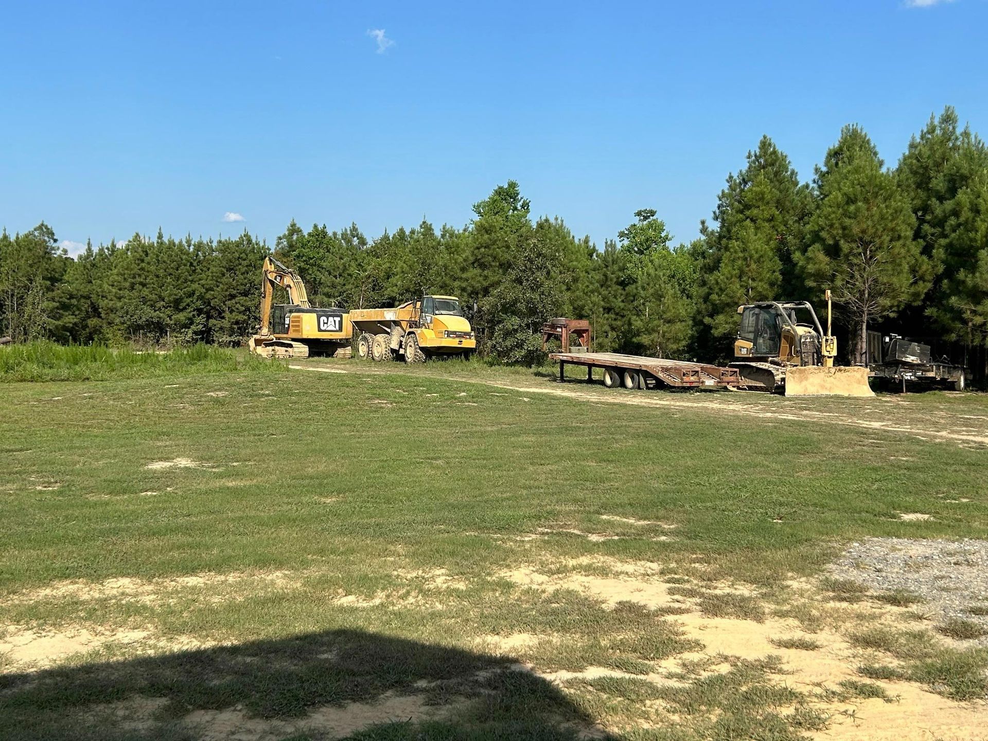 Yellow construction equipment on a grassy field with trees in the background under a blue sky.