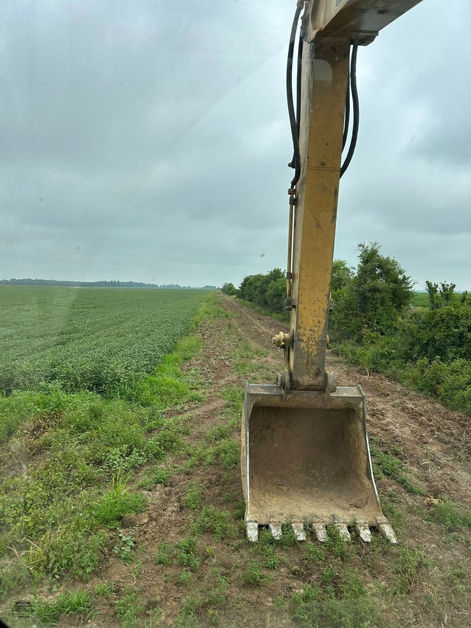 Yellow excavator bucket next to a field with green crops and a cloudy sky.