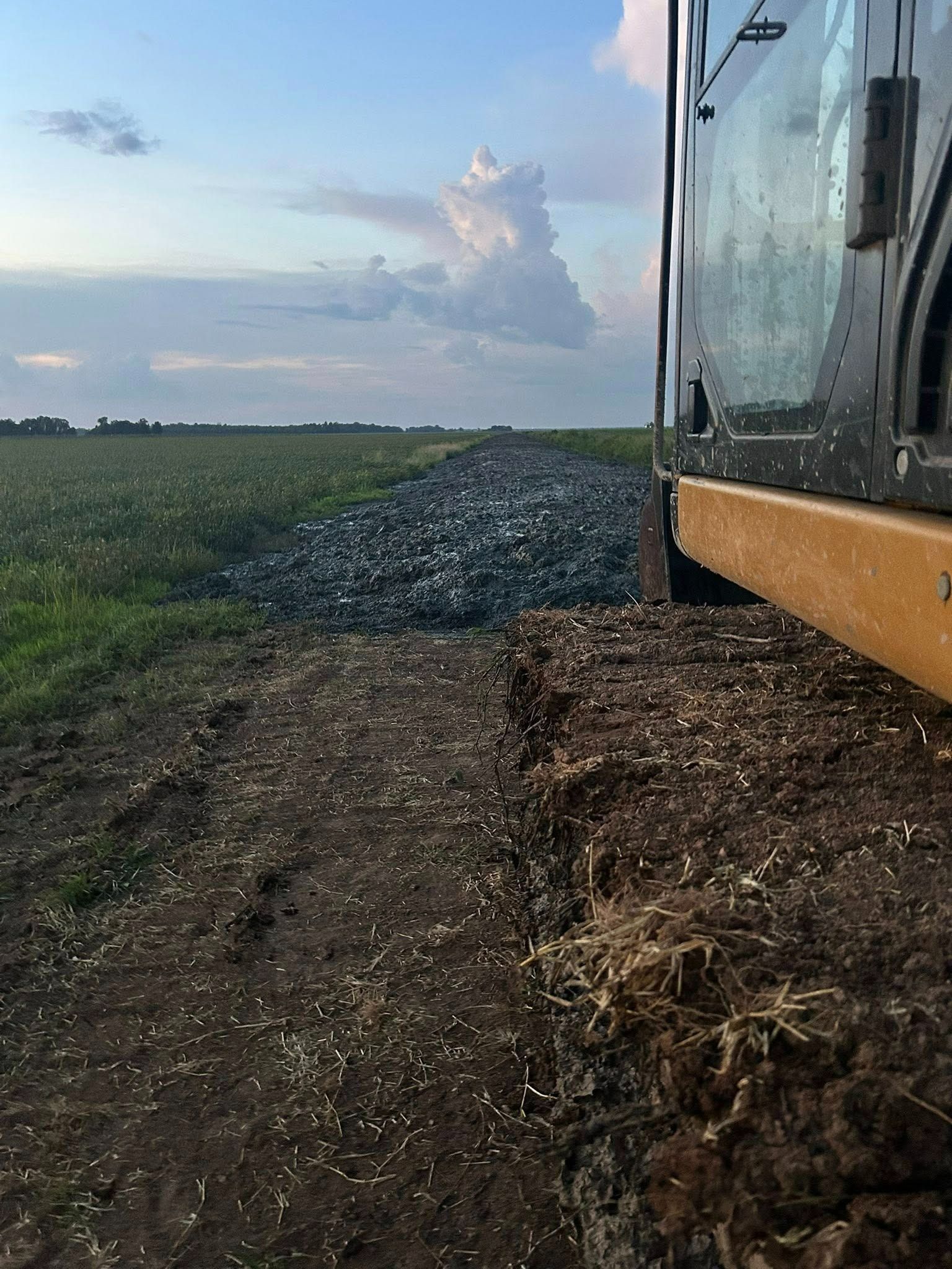 A construction vehicle on a dirt path next to a gravel road, with a field and cloudy sky in the background.