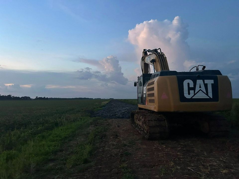 A yellow Caterpillar excavator parked on a dirt path next to a pile of rocks in a field, under a cloudy sky.