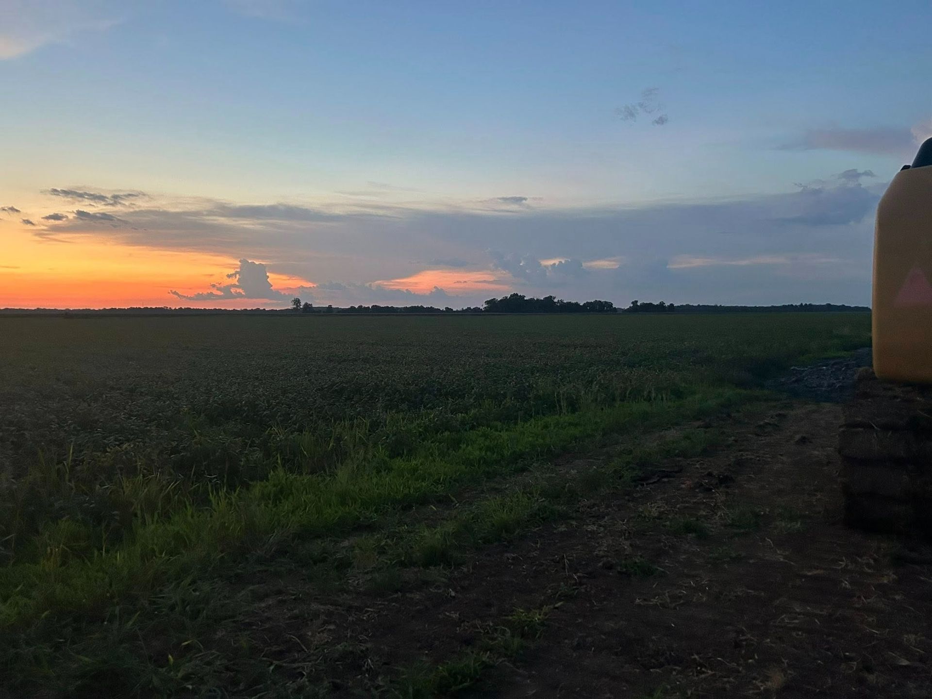 Sunset over a field; orange and blue sky, dark green plants, and a glimpse of machinery.