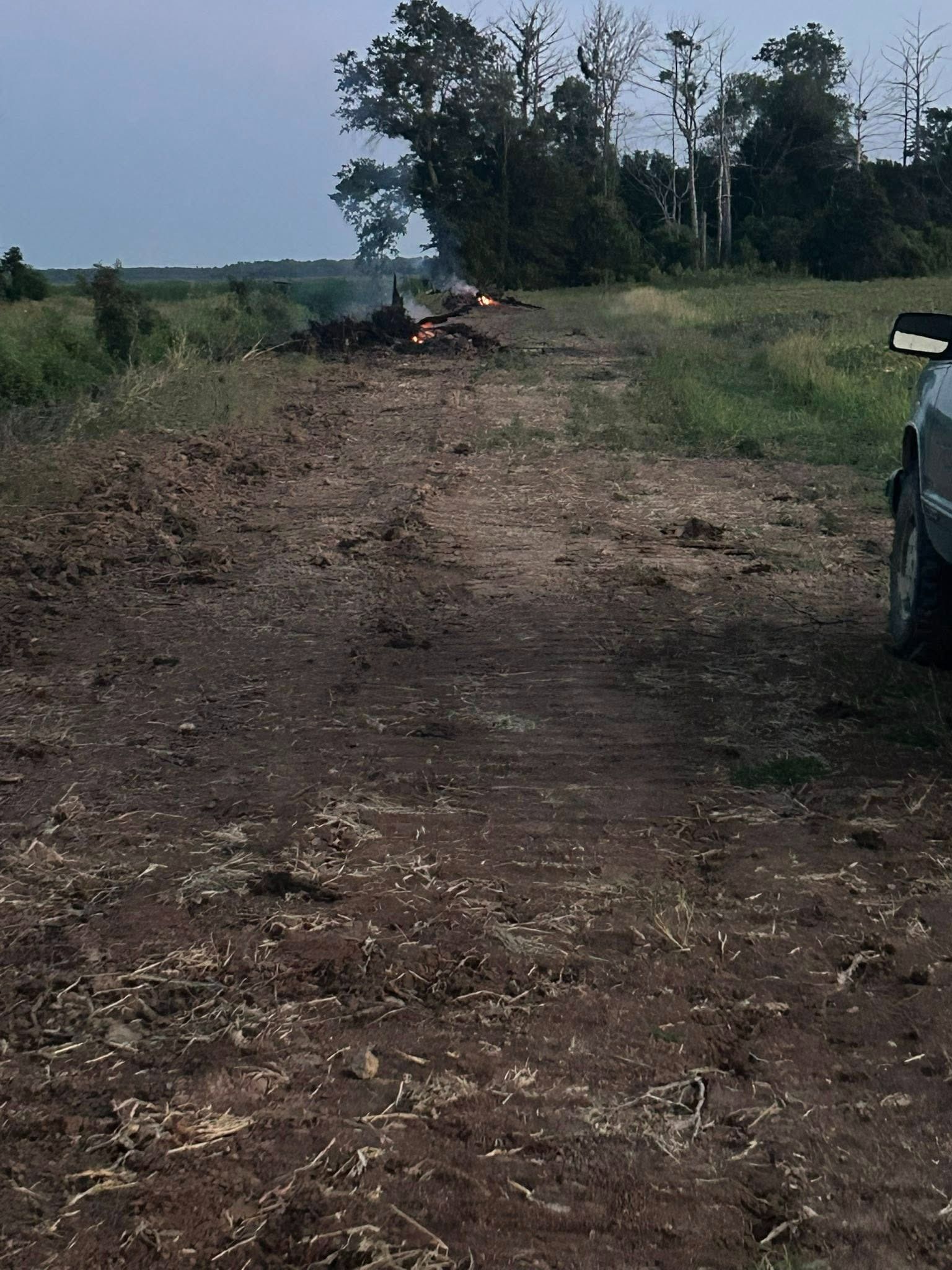 Dirt road leading to a brush fire in a field at dusk, with a vehicle parked on the right.