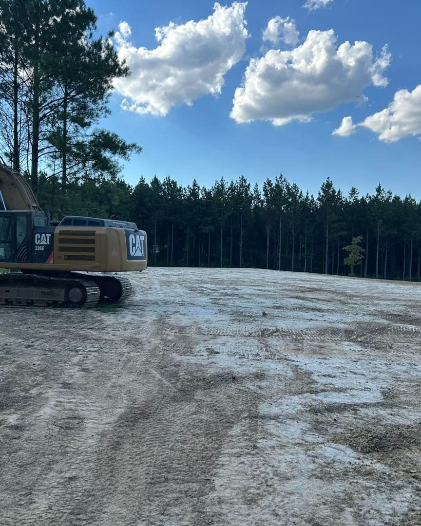 A cleared dirt lot with a construction excavator, trees in the background, and a blue sky.