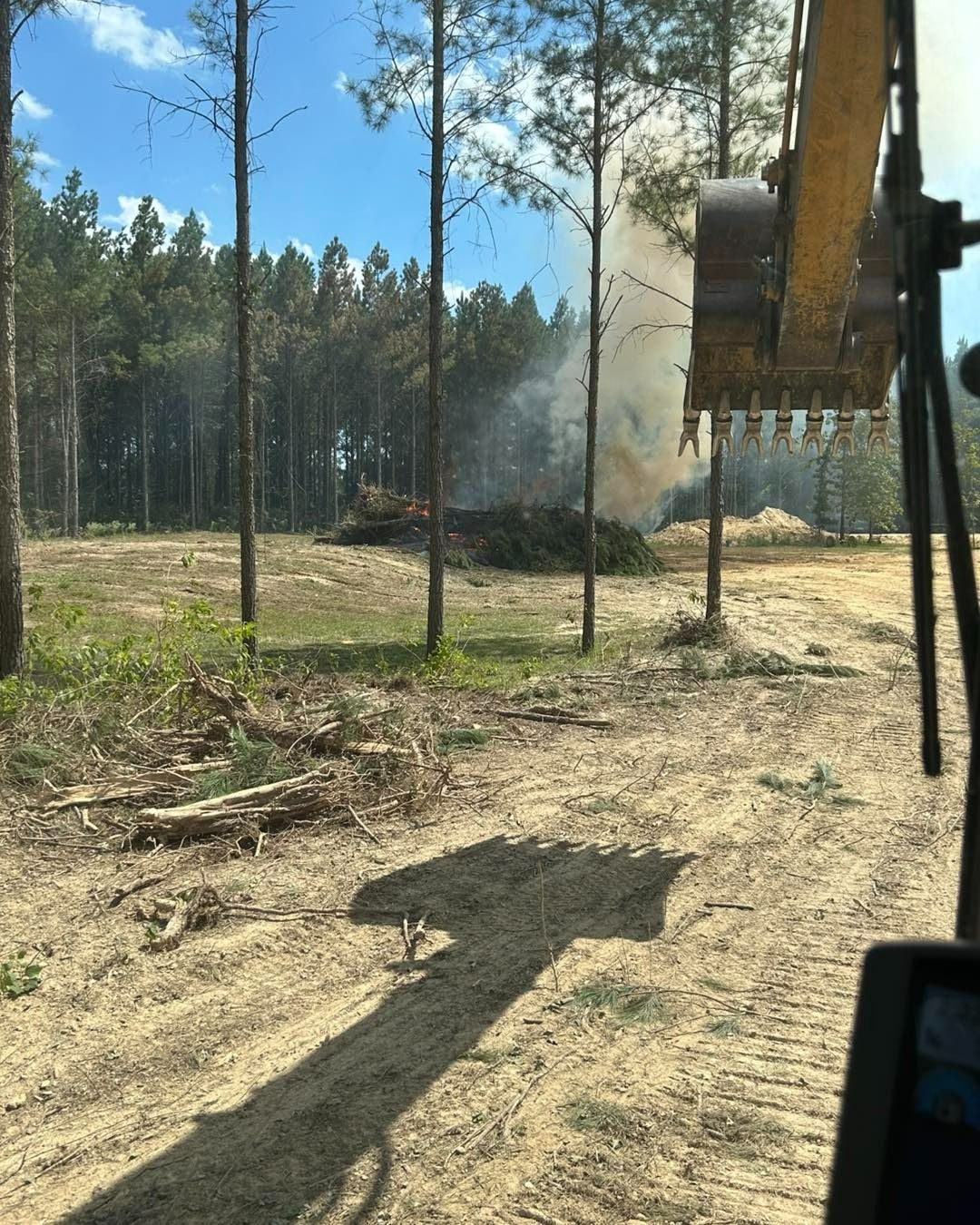 An excavator with debris burning in a clearing. Pine forest in the background, sunny day.