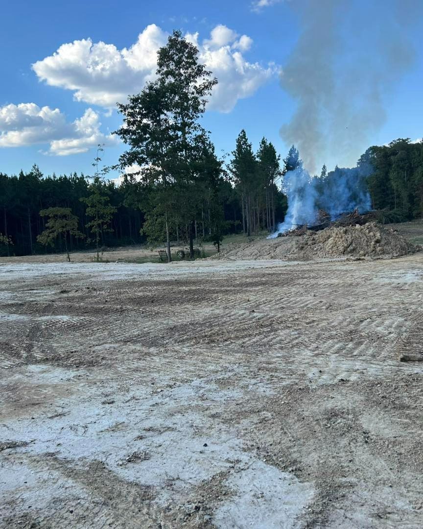 Smoke rising from a debris pile in a cleared field, trees in the background, under a partly cloudy sky.