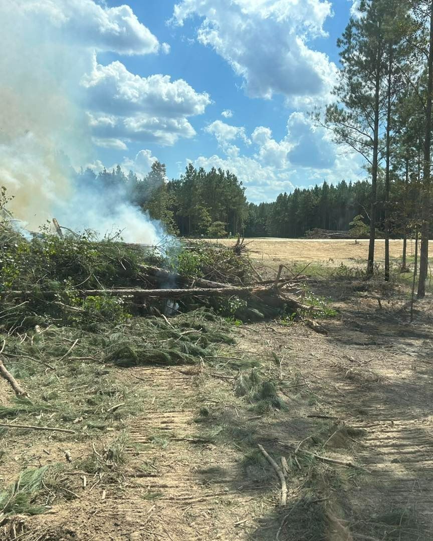 Smoke rising from a brush pile burning in a field, with trees in the background and a partly cloudy sky.