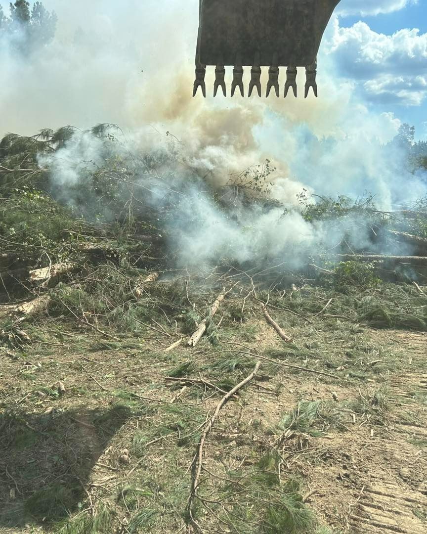 Excavator over a burning pile of brush, producing smoke in a field.