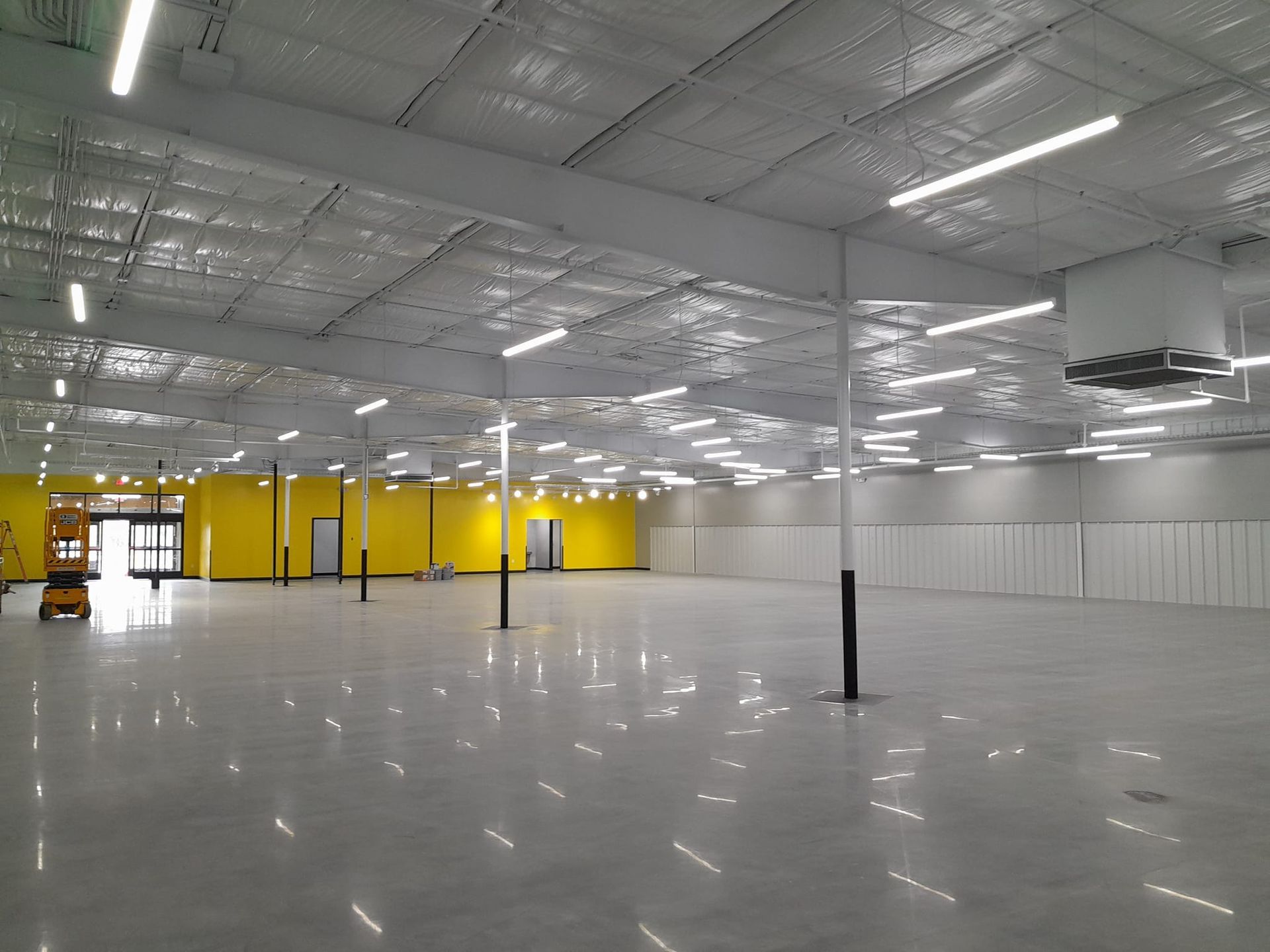 A wide-open, industrial warehouse interior with a reflective concrete floor, bright yellow walls, and high ceiling lights.