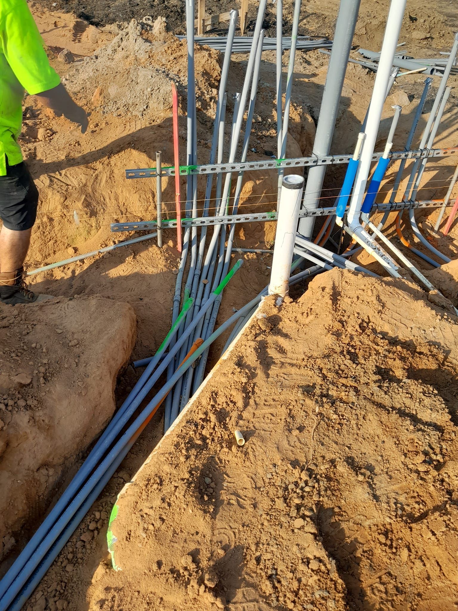 A person in a high-visibility shirt stands by a trench filled with multiple electrical conduits and metal supports.