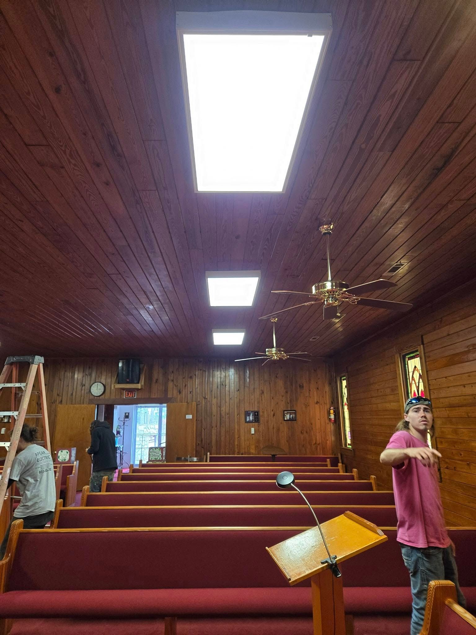 A church interior with wooden walls, rows of red pews, a podium, and ceiling lights, with two people in the room.