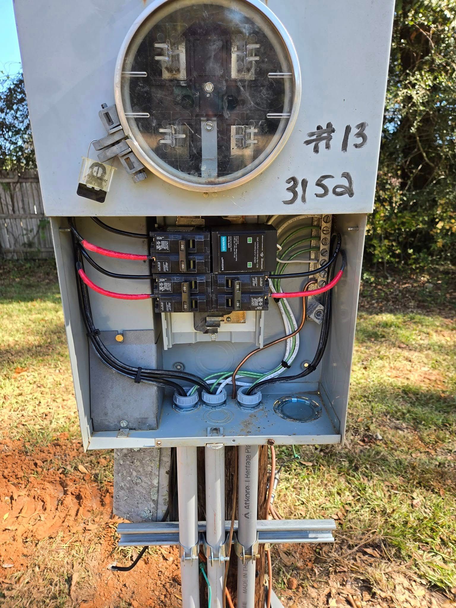 An open electrical meter box mounted on a pole outdoors, showing internal wiring, two breakers, and conduits below.