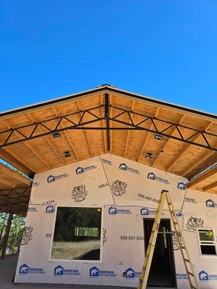 The front exterior of a house under construction with exposed wooden roof trusses, plywood sheathing, and a ladder.