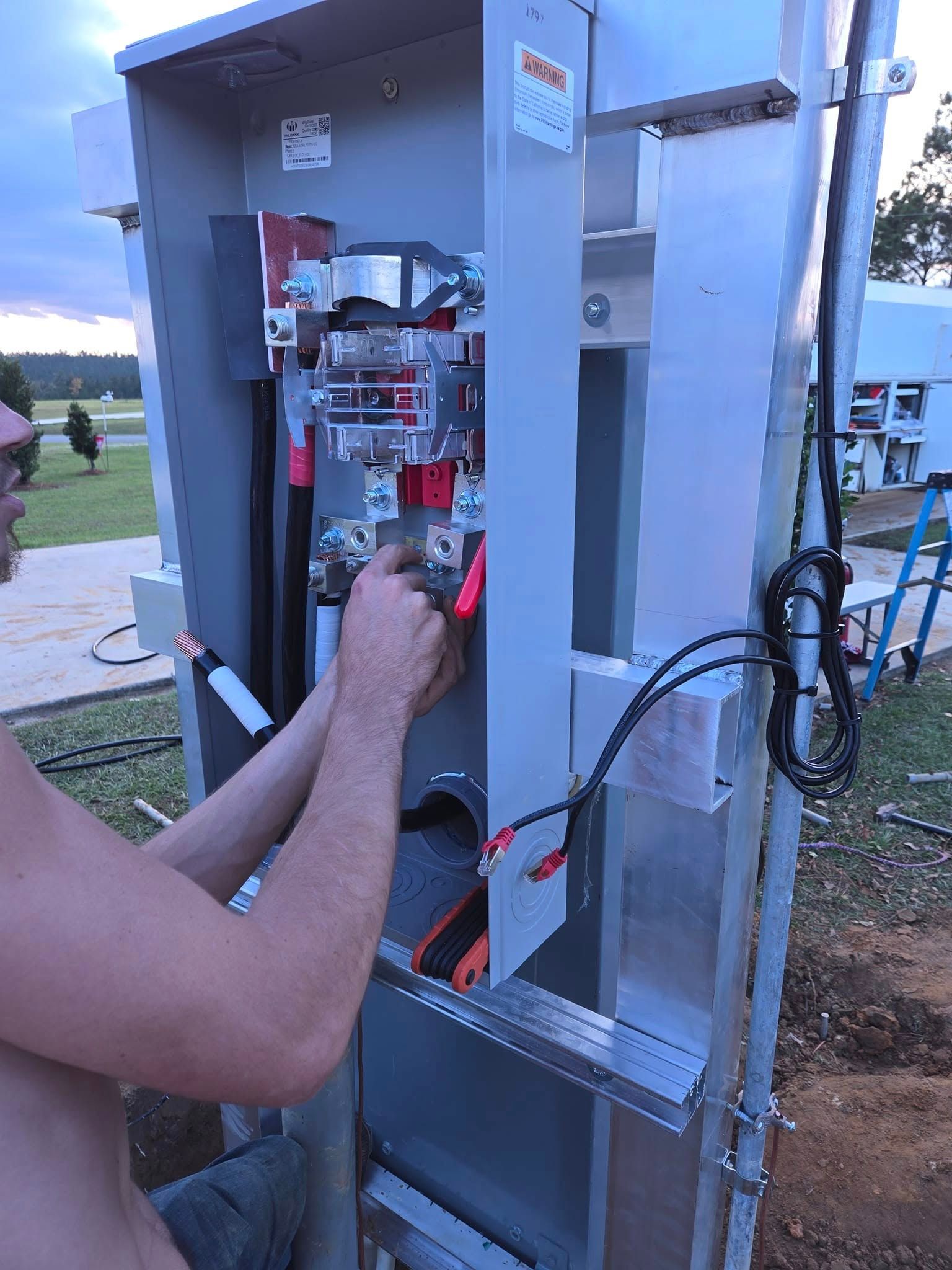 A person works on the wiring inside an open outdoor electrical meter box set on a metal frame.