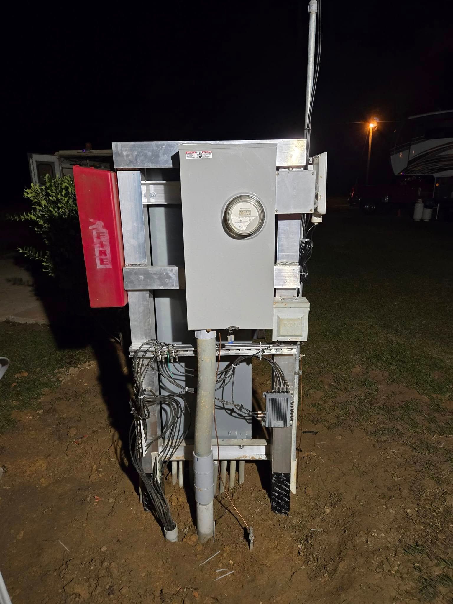 An electrical meter and control box mounted on a metal frame at night, set in an outdoor, grassy area.