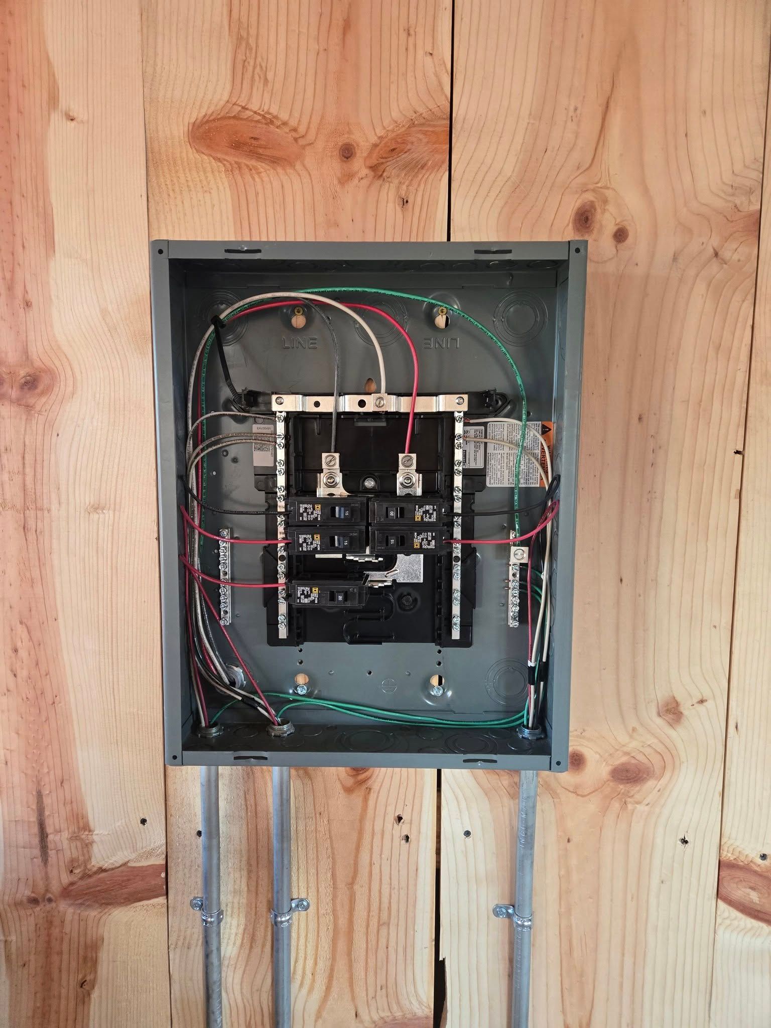 An open electrical service panel mounted on a wood wall, showing wiring connected to breakers inside a gray metal box.