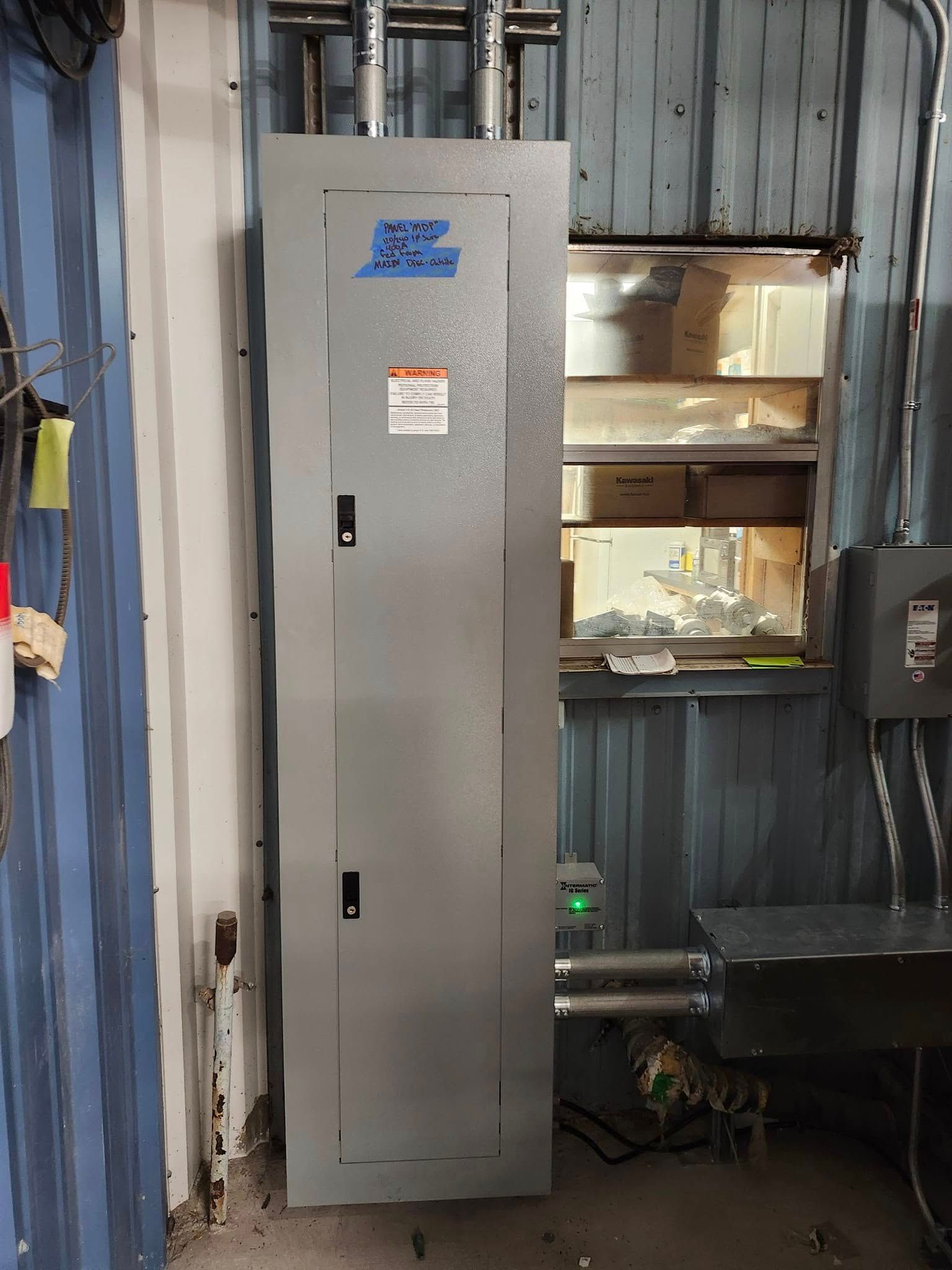 A gray industrial electrical panel mounted on a blue metal wall, with conduit pipes leading to a nearby junction box.