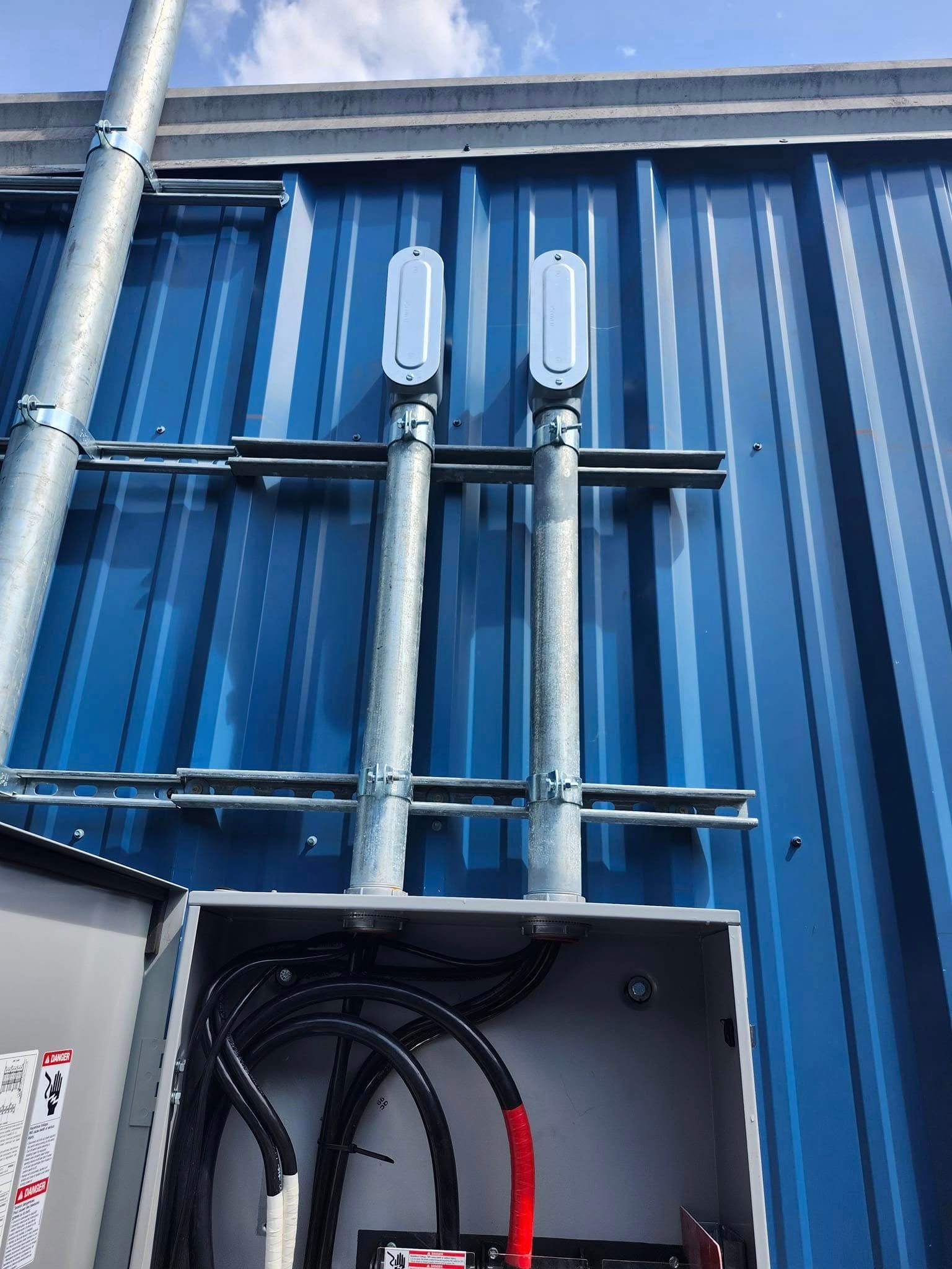 Two metal electrical conduits with access covers mounted on a blue corrugated metal wall above an open junction box.