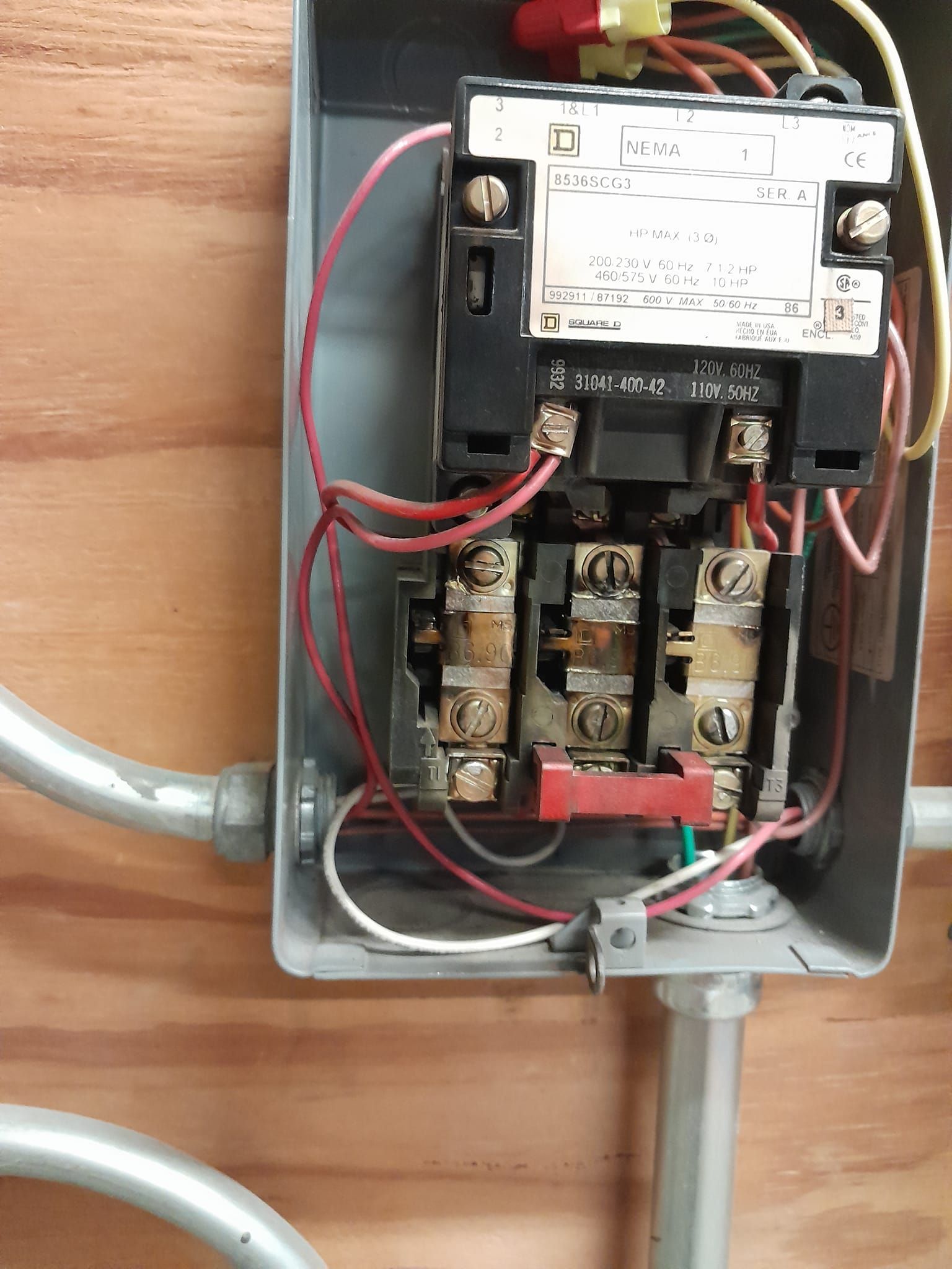 An electrical contactor mounted on a wooden wall, featuring black plastic casing, metal screw terminals, and red wiring.
