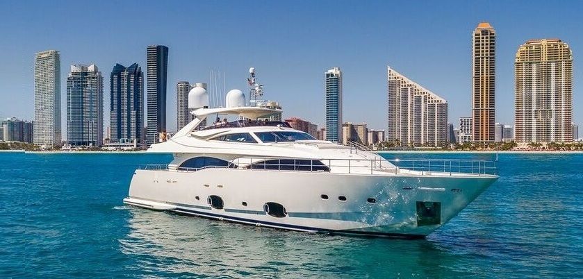 A luxury yacht floats in front of a cityscape with tall buildings on a sunny day.