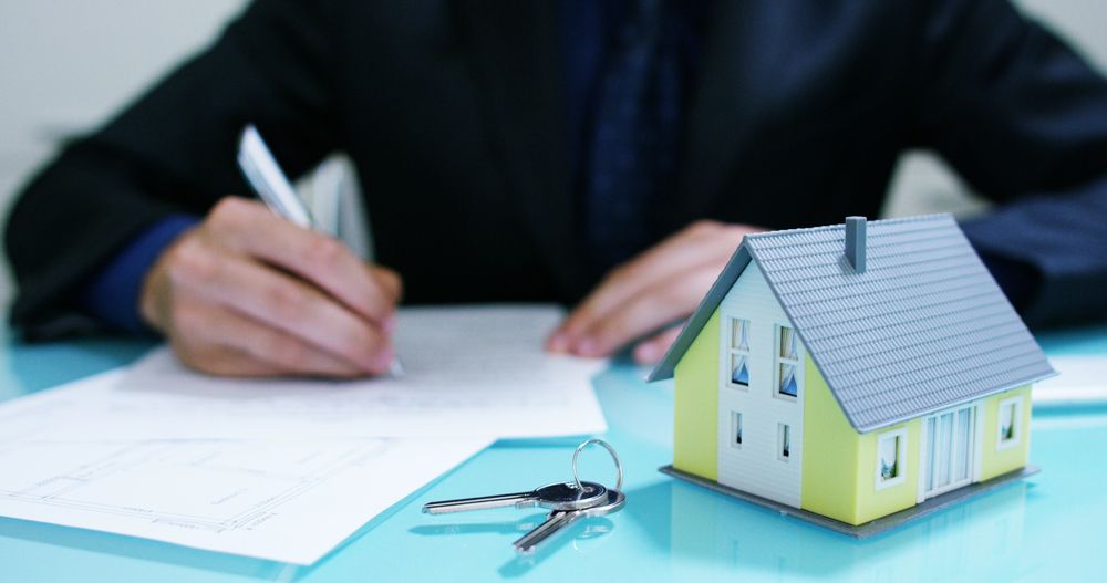 A Man is Writing on a Piece of Paper Next to a Model House and Keys — Karlene's Conveyancing In Avoca, NSW