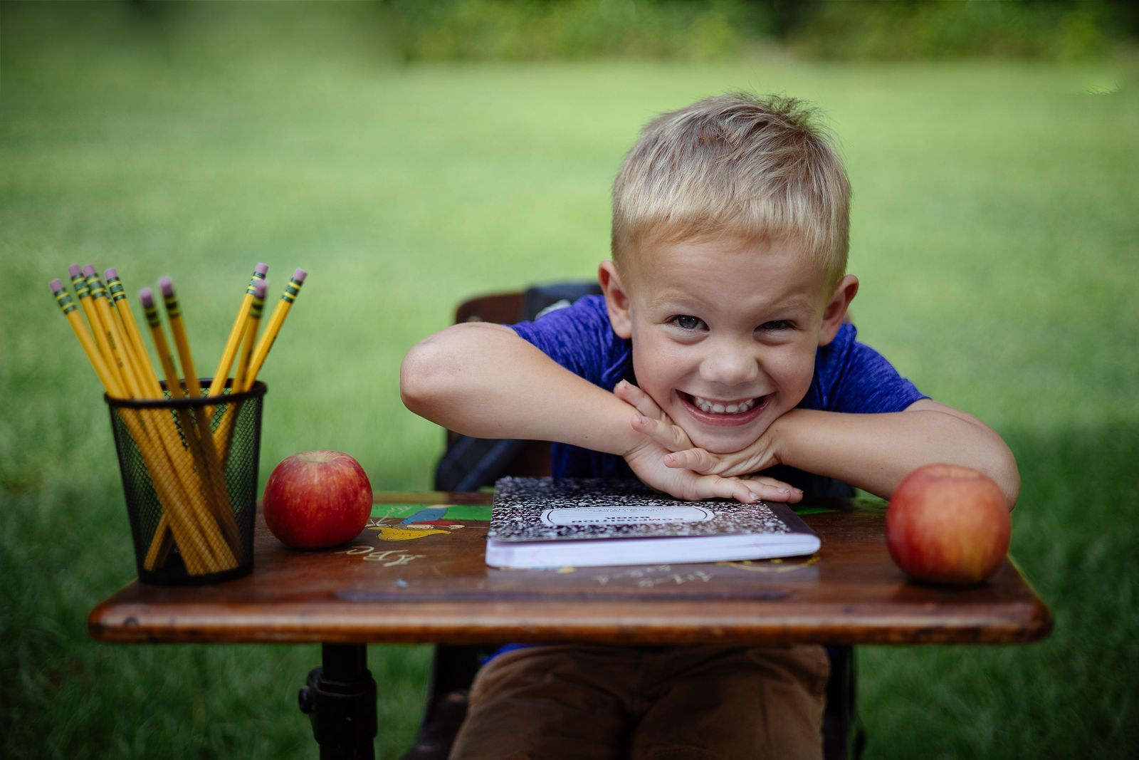 Boy with blonde hair smiles at desk with pencils, notebook, and apples. Green background.