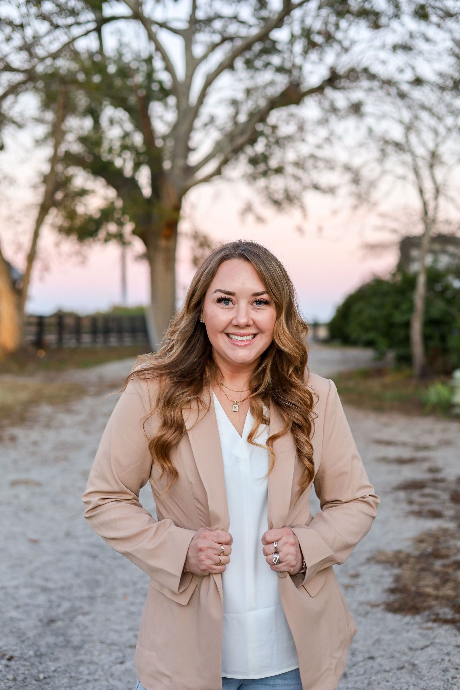 Woman in tan blazer smiles outdoors, hands on jacket. Tree and blurred background.