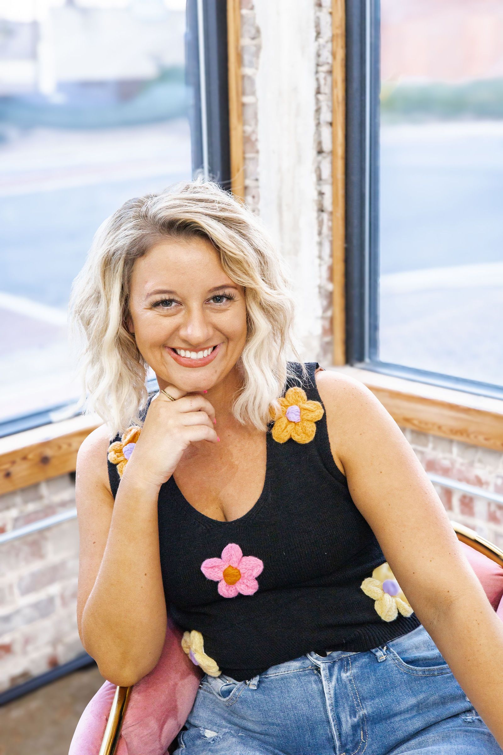 Woman with curly blonde hair smiles, wearing a black top with flower accents and acid wash jeans, seated by a window.