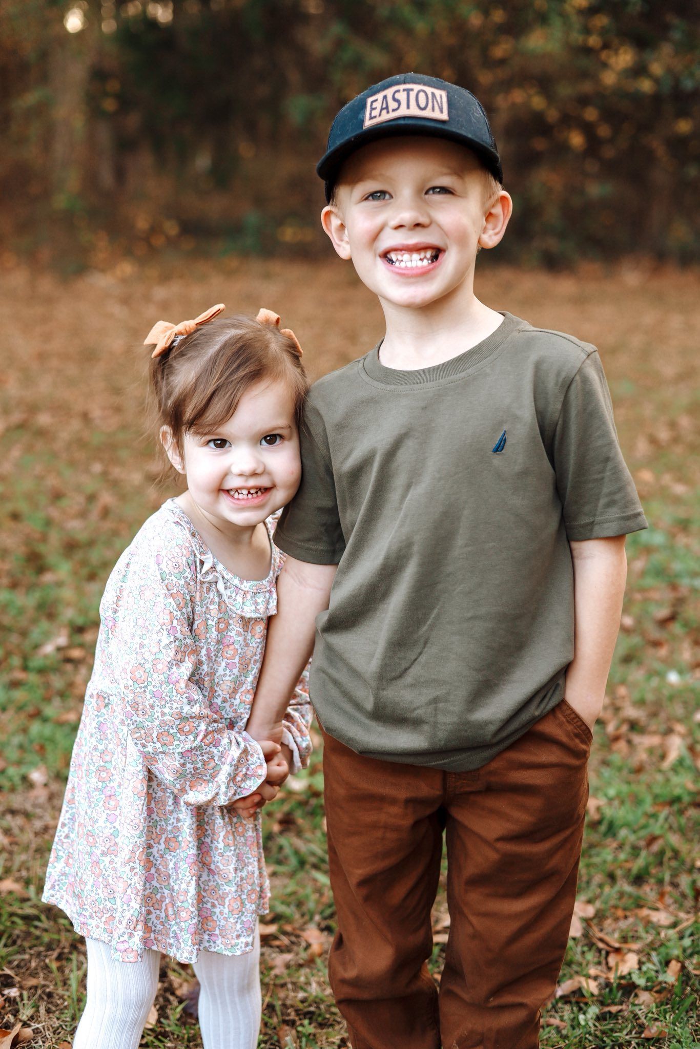 Girl and boy holding hands, smiling. Girl in floral dress, boy in olive shirt and hat, standing outside.