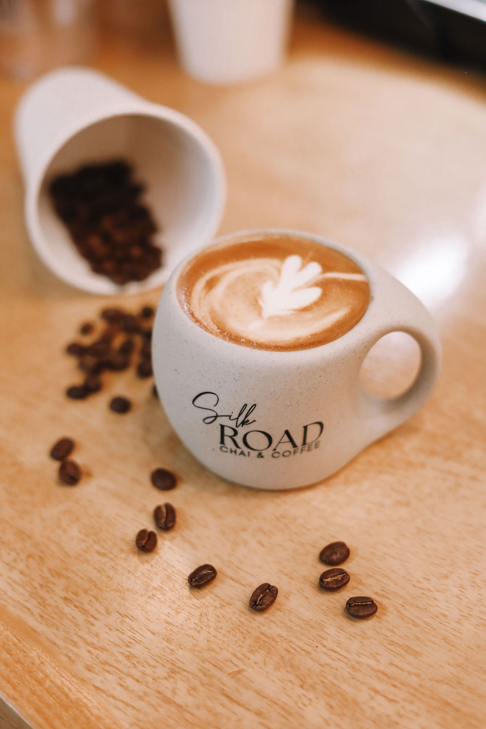 Latte in a speckled mug with leaf art, spilled coffee beans and cup on a wooden table at a cafe. strategic branding shoot for coffee company 