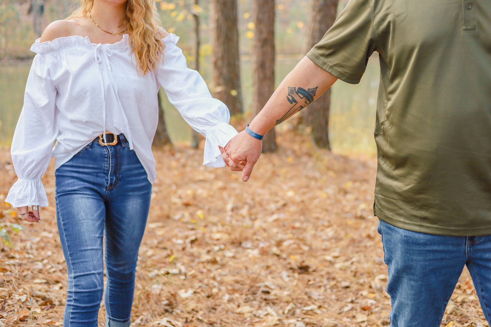 Couple holding hands, walking in a wooded area with fall foliage. The woman wears a white top and jeans. The man wears a green shirt and jeans.